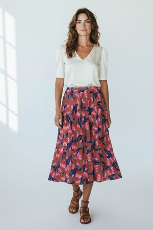 A woman with wavy hair smiles softly in a bright room, wearing a white V-neck blouse, brown strappy sandals, and the FRANCO FERRARI - FLORAL PRINT SKIRT—a high-waisted pink and navy made-in-Italy piece by FRANCO FERRARI.