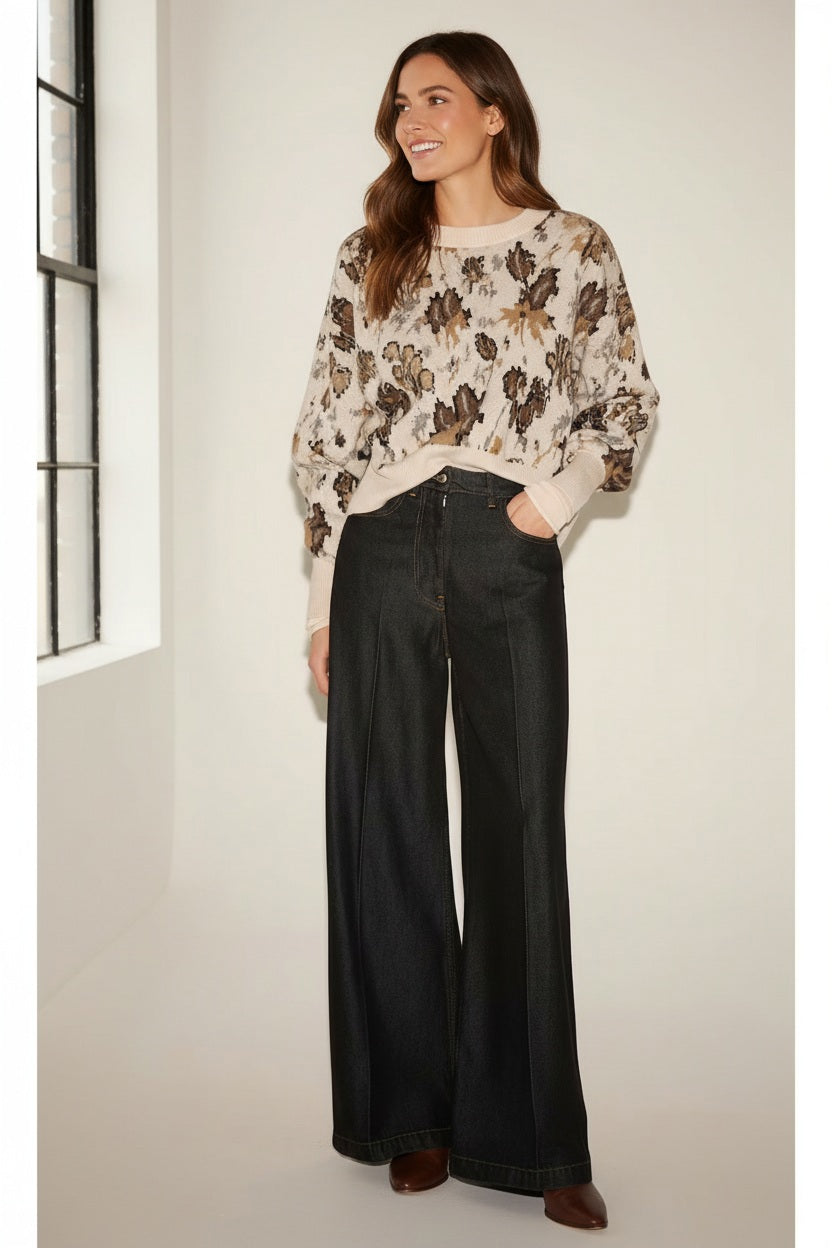 A woman with long brown hair stands indoors near a window, smiling and looking to the side. She is wearing a beige floral long-sleeve sweater paired with the NUDE TILDA Wide Leg Trouser in Black by NUDE. One hand is tucked into her pocket.