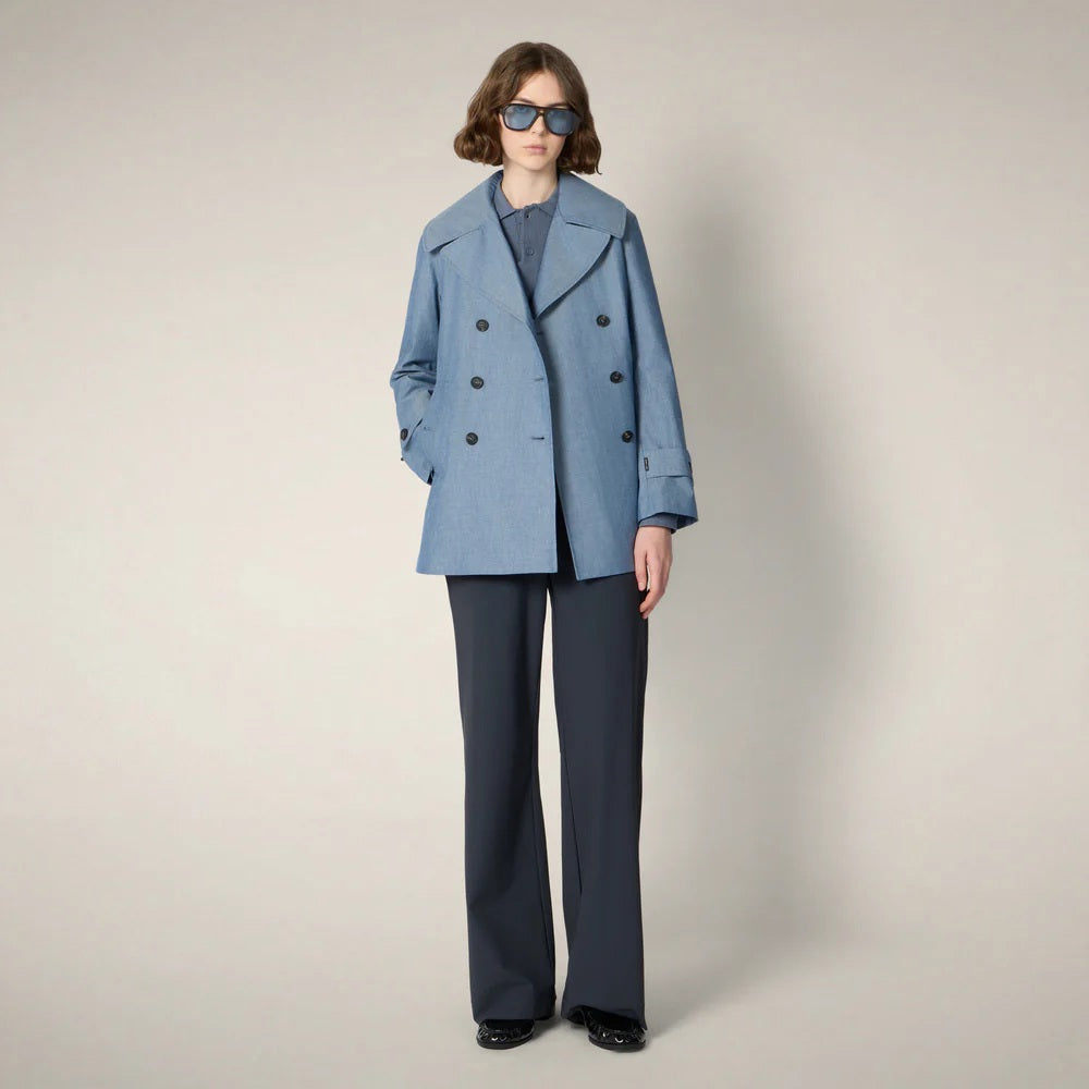 A person wearing dark sunglasses, the SAVE THE DUCK SELA Peacoat Raincoat in Chambray, and wide-leg dark pants stands against a plain light background.