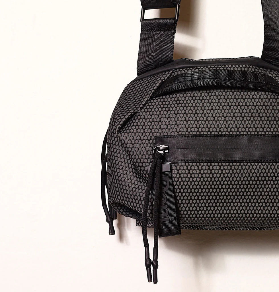 A person in a light beige suit carries the DANIELLA LEHAVI - BONNIE TRAVEL CROSSBODY, a large brown vegan bag with black zipper and strap, shown from shoulders to mid-thigh against a plain background.