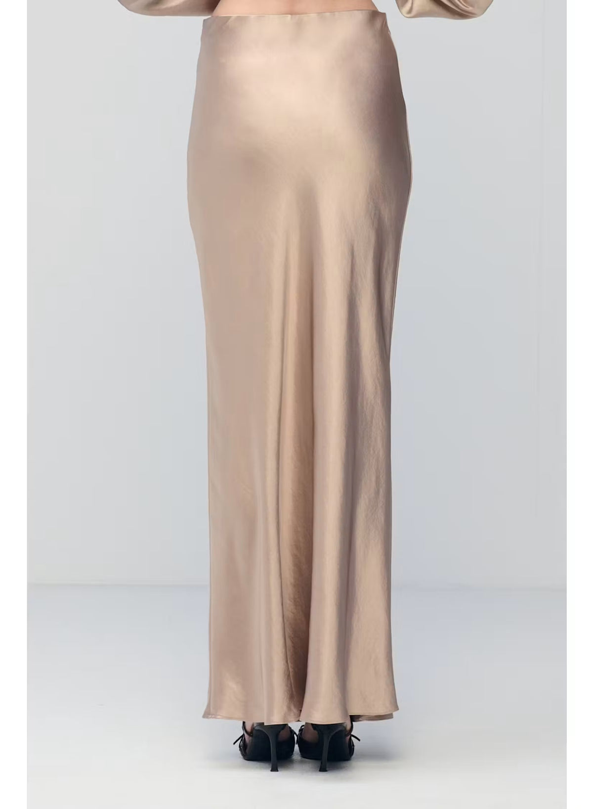 A person wearing the MILVA MI - LONG SATIN SKIRT IN SAND by MILVA, paired with pointed black shoes and standing against a plain, light background.