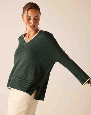 A person wearing the MERSEA Montauk Colorblock V Neck Sweater in Eden, paired with off-white pants and brown ankle boots, stands in a sunlit room with white walls and light wood floors.