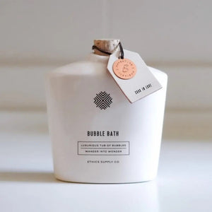 The elegant white "SOAK IN LOVE" ceramic bubble bath bottle from ETHICS SUPPLY CO, complete with a cork stopper, sits on rustic wood. Nearby, a natural sea sponge is surrounded by lavender sprigs and the scent of essential oils, while a window blurs green scenery outside.