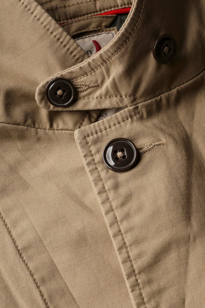 A person wears the RELWEN Flyweight Flex Blazer in British Khaki over a blue and white checkered shirt with green pants, standing hands-in-pockets against a white background. Face not visible.