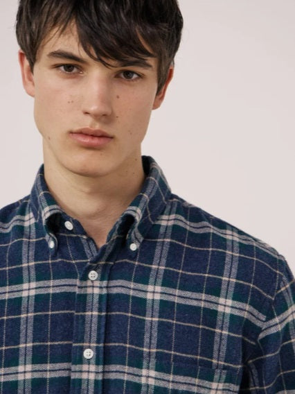 A young person with dark hair, wearing the HARTFORD - PITT CHECKED FLANNEL SHIRT IN BLUE AND GREEN by HARTFORD, looks at the camera while adjusting the shirt collar with one hand against a plain light background.