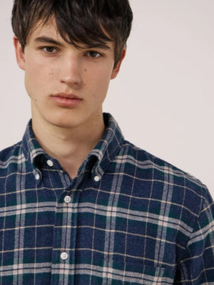 A young person with dark hair, wearing the HARTFORD - PITT CHECKED FLANNEL SHIRT IN BLUE AND GREEN by HARTFORD, looks at the camera while adjusting the shirt collar with one hand against a plain light background.