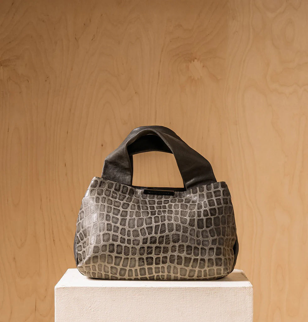 The DANIELLA LEHAVI Eveline Satchel in Grey Mosaic features a croco-inspired textured leather design with short handles and a long shoulder strap, epitomizing quiet luxury as it hangs gracefully against a wooden background.
