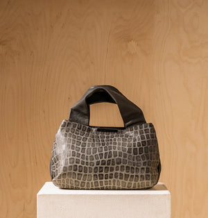 The DANIELLA LEHAVI Eveline Satchel in Grey Mosaic features a croco-inspired textured leather design with short handles and a long shoulder strap, epitomizing quiet luxury as it hangs gracefully against a wooden background.