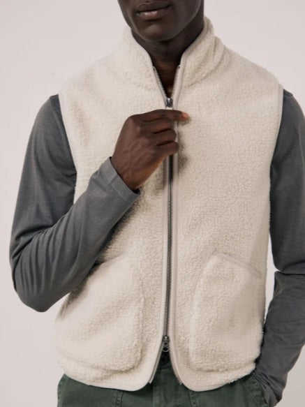 A man is wearing the HARTFORD - BASTA SHERPA VEST IN NATURAL over a gray long-sleeve shirt, holding the vest’s zipper with one hand. The plain, light-colored background complements the cozy, Hartford-inspired style of this men’s vest from HARTFORD.