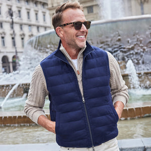 A smiling man in a light blue shirt and the COLLARS AND CO - REGENT REVERSIBLE QUILTED WOOL VEST holds a blue umbrella on a rainy street—showcasing COLLARS AND CO's ideal insulated layering for wet days.