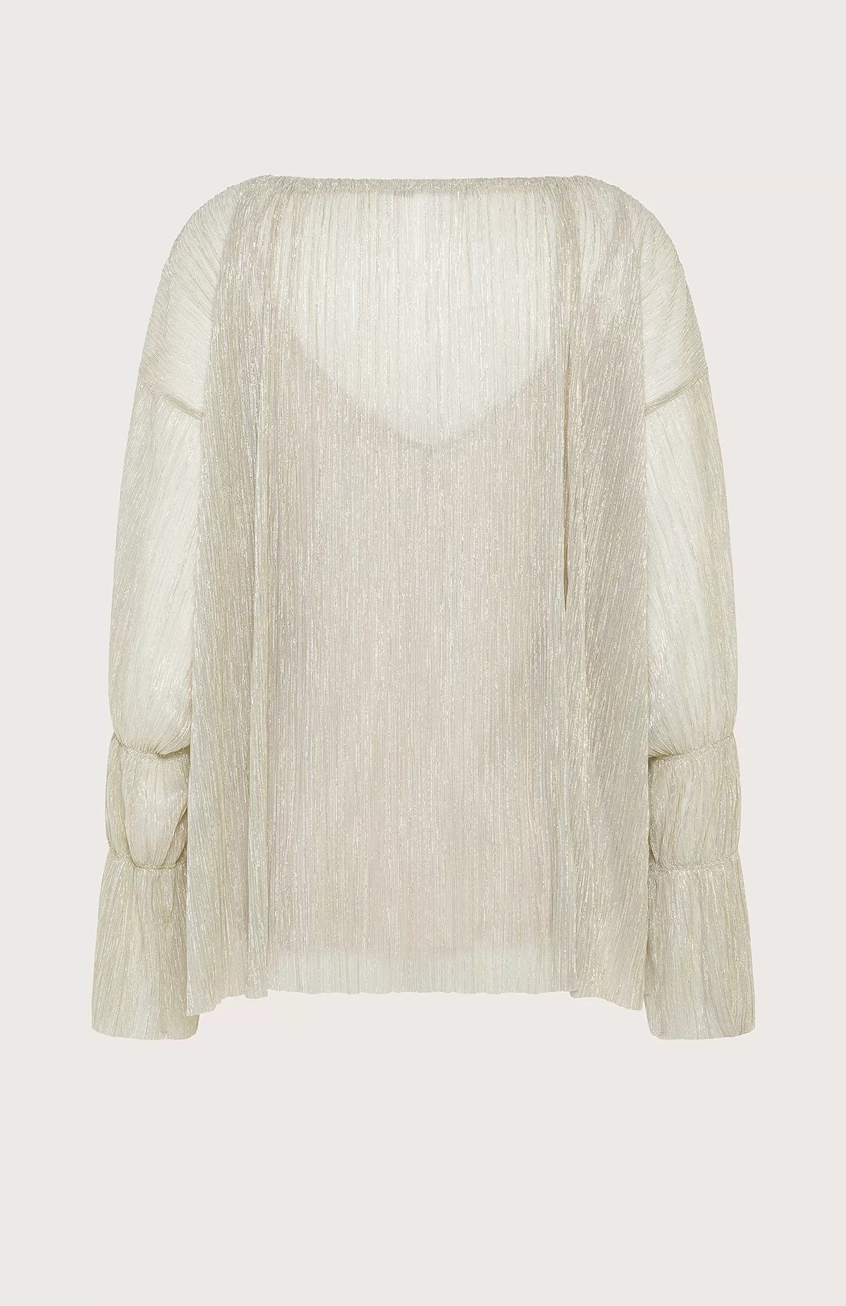 A person with shoulder-length hair stands against a light blue background, wearing the SEVENTY VENEZIA LUREX JERSEY BLOUSE by SEVENTY, which is a shimmery beige pleated blouse with long sleeves, paired with wide-leg beige trousers. The person gazes forward with a relaxed posture, one hand in their pocket.