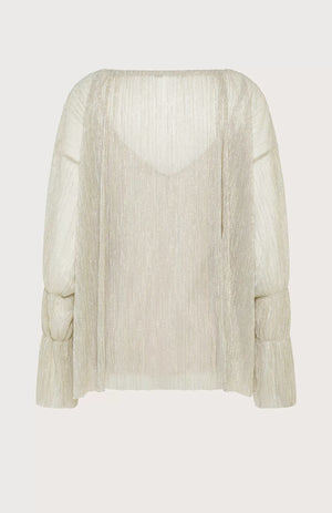 A person with shoulder-length hair stands against a light blue background, wearing the SEVENTY VENEZIA LUREX JERSEY BLOUSE by SEVENTY, which is a shimmery beige pleated blouse with long sleeves, paired with wide-leg beige trousers. The person gazes forward with a relaxed posture, one hand in their pocket.