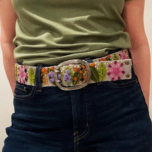 A person wears a magenta long-sleeve top, dark blue jeans, and the JENNY KRAUSS - FRESHWATER BELT by JENNY KRAUSS, handcrafted in Peru and adorned with colorful embroidered flowers and leaves.