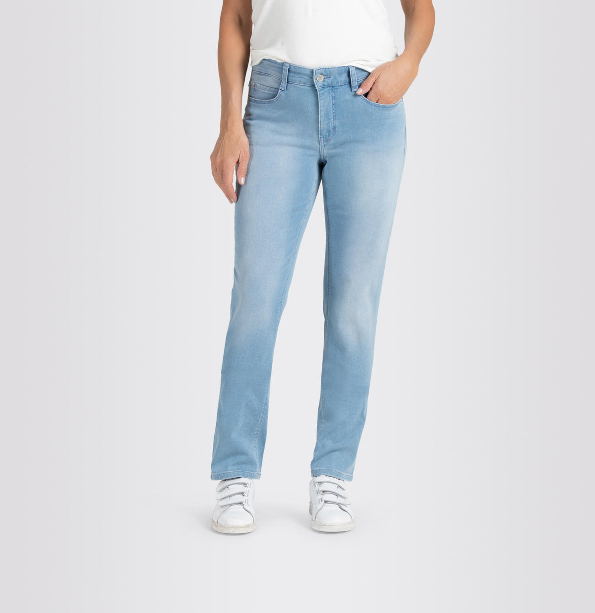 A person is standing against a plain background wearing MAC&#39;s DREAM STRAIGHT LEG DENIM jeans in light blue, paired with a white shirt and white sneakers. Their right hand is in their pocket while their left hand hangs down by their side. The slim fit of the MAC jeans provides a subtle shaping effect for a tailored look.