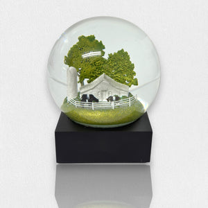 The FEARRINGTON SIGNATURE COLLECTION - FEARRINGTON BELTIE SNOWGLOBE from COOL SNOW GLOBES features a charming miniature rural scene complete with a white farmhouse, black cows, a tall silo, and green trees, all enclosed by a white picket fence. This delightful piece rests on a black square base against a white background.