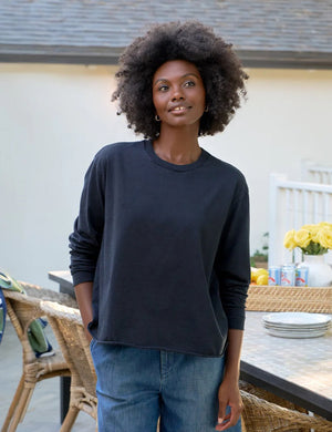 A woman with naturally curly hair, wearing the FRANK & EILEEN QUINN Long Sleeve Crewneck Tee in British Royal Navy and relaxed blue jeans, stands by an outdoor table with plates, drinks, and flowers, smiling softly. Made in California.