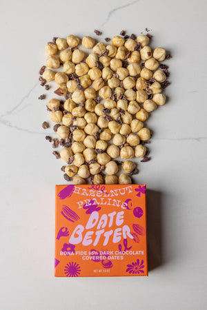 The "DATE BETTER - HAZELNUT PRALINE COVERED DATES" from DATE BETTER SNACKS features orange packaging with bold pink and purple text, adorned with abstract flower and leaf patterns. This 85% dark chocolate Medjool date treat weighs 1.8 oz.