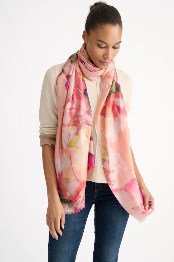 A woman wearing a light sweater and jeans stands with the KINROSS CASHMERE - HIGHLAND BLOOMS SCARF IN SANDSTONE from KINROSS draped around her neck, looking down with a gentle expression against a plain white background.
