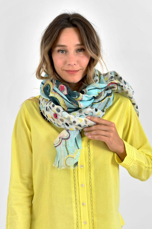 A woman in a yellow sweater and beige skirt holds up the STORIATIPIC - ANNE SCARF IN AQUA, a colorful linen scarf by STORIATIPIC featuring floral and polka dot patterns with exquisite handmade embroidery, against a white background.