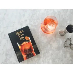 The SHAKE. STIR. SIP. BY KARA NEWMAN cocktail book by CHRONICLE BOOKS features a cover with an amber drink garnished with orange peel and white text: "Shake. Stir. Sip." plus "More than 50 simple cocktails made in equal parts.