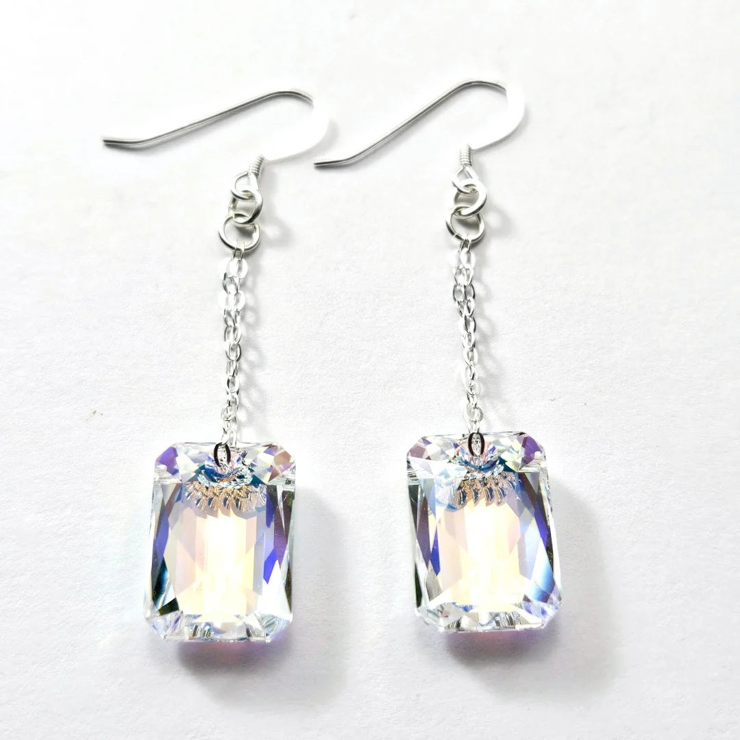 The VERDIER - SWAROVSKI EMERALD CUT CRYSTAL EARRINGS by VERDIER feature sterling silver hooks and rectangular, emerald cut crystals inspired by the Swarovski Aurora design, delicately suspended from fine chains and elegantly presented against a white background.