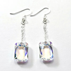 The VERDIER - SWAROVSKI EMERALD CUT CRYSTAL EARRINGS by VERDIER feature sterling silver hooks and rectangular, emerald cut crystals inspired by the Swarovski Aurora design, delicately suspended from fine chains and elegantly presented against a white background.