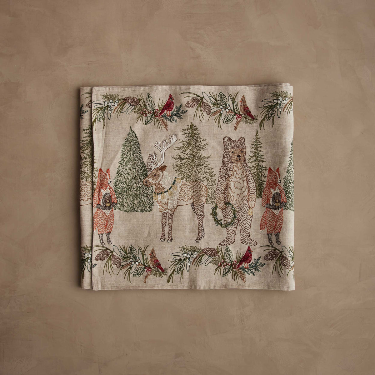 A CHRISTMAS CHEER EMBROIDERED LINEN TABLE RUNNER featuring woodland animals in winter attire and Christmas trees, draped over a wooden dining table set with plates, glassware, and candlesticks.