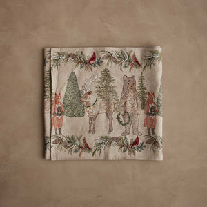 A CHRISTMAS CHEER EMBROIDERED LINEN TABLE RUNNER featuring woodland animals in winter attire and Christmas trees, draped over a wooden dining table set with plates, glassware, and candlesticks.