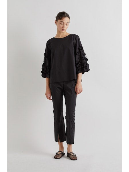A person stands against a plain background wearing MEIMEIJ FROTN HEM SPLIT COTTON PANT—black, cropped, ankle-length pants with small slits. They also wear a black textured-sleeve top and black shoes. Their face is not visible in the image.