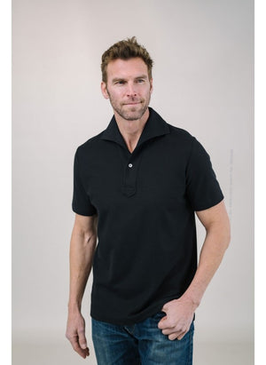 A man with short brown hair and a trimmed beard models the EDWARD ARMAH Pima Cotton Tailored Polo Shirt in white with light-colored pants, standing against a plain light background, one hand in his pocket, looking to the side.