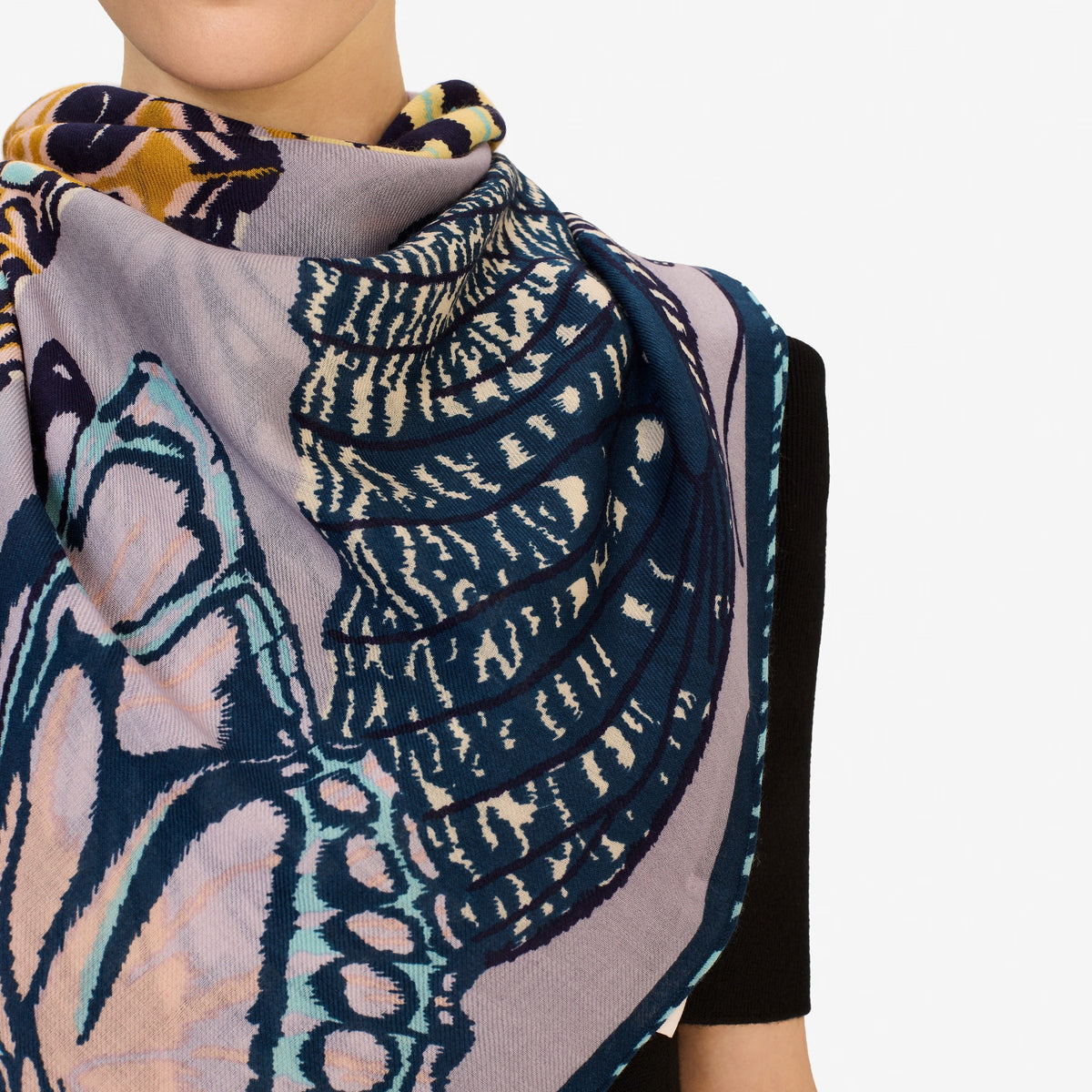 A woman with straight brown hair pulled back wears a dark top and drapes the INOUI EDITIONS PAPILLON (BUTTERFLIES) Square Scarf in Grey by INOUI, featuring abstract and butterfly patterns, around her neck against a white background.
