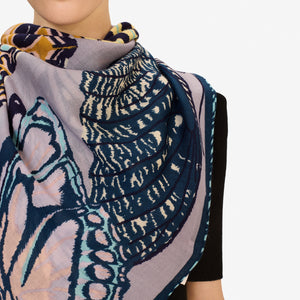 A woman with straight brown hair pulled back wears a dark top and drapes the INOUI EDITIONS PAPILLON (BUTTERFLIES) Square Scarf in Grey by INOUI, featuring abstract and butterfly patterns, around her neck against a white background.