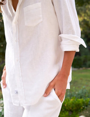 A woman with curly hair smiles outdoors, wearing the FRANK & EILEEN - EILEEN Relaxed Button Up Shirt in linen from the Tuscany Getaway Set, paired with wide-leg pants, standing on a sunlit stone patio surrounded by greenery.