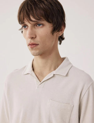 Wearing the HARTFORD Terry Polo Shirt in Stone with sage green cargo pants, a person stands with one hand in their pocket, glancing to the side against a plain background.