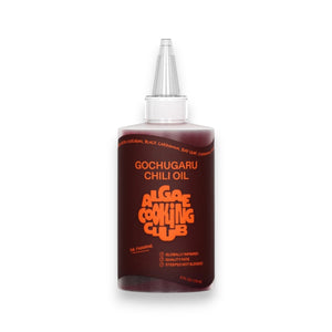A clear plastic bottle labeled "ALGAE COOKING CLUB - GOCHUGARU CHILI OIL" in bold orange text sits on a white surface against a plain background, holding vibrant Korean chili oil from ALGAE COOKING CLUB.