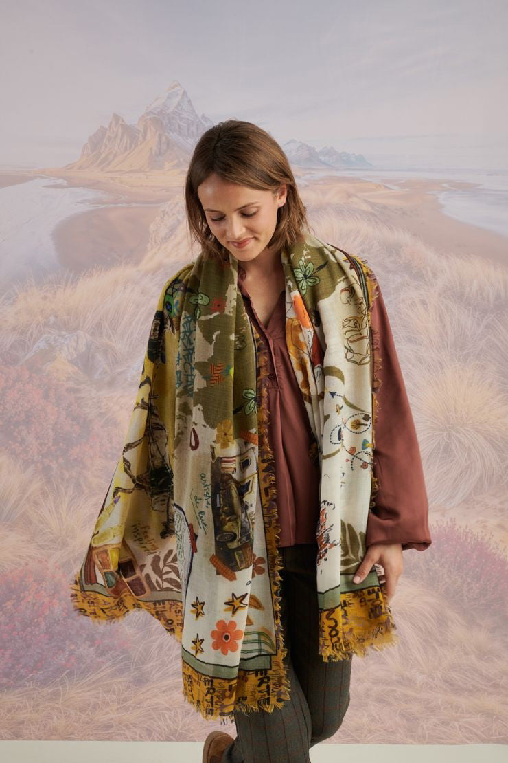 A woman with wavy blonde hair smiles while looking down. She wears a loose, light lavender blouse, dark pants, and the STORIATIPIC EUGENIE WOOL SCARF. Behind her is a scenic landscape of mountains, grass, and water.