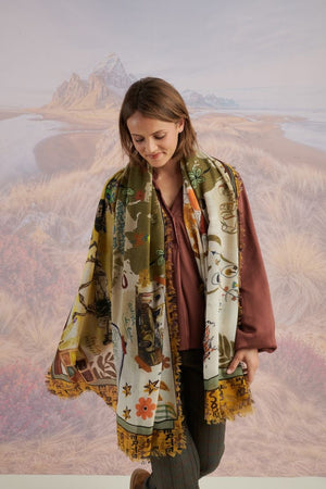 A woman with wavy blonde hair smiles while looking down. She wears a loose, light lavender blouse, dark pants, and the STORIATIPIC EUGENIE WOOL SCARF. Behind her is a scenic landscape of mountains, grass, and water.