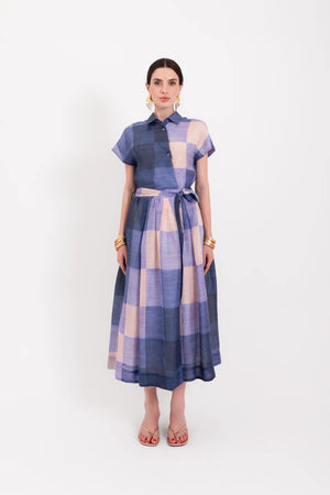 A woman wearing the LAVI - BELLELL Dress in Plaid by LAVI, a short-sleeve, collared shirt dress with a blue and beige plaid pattern and a tied waist, accessorized with gold earrings, bracelets, and open-toed heels, stands against a plain white background.