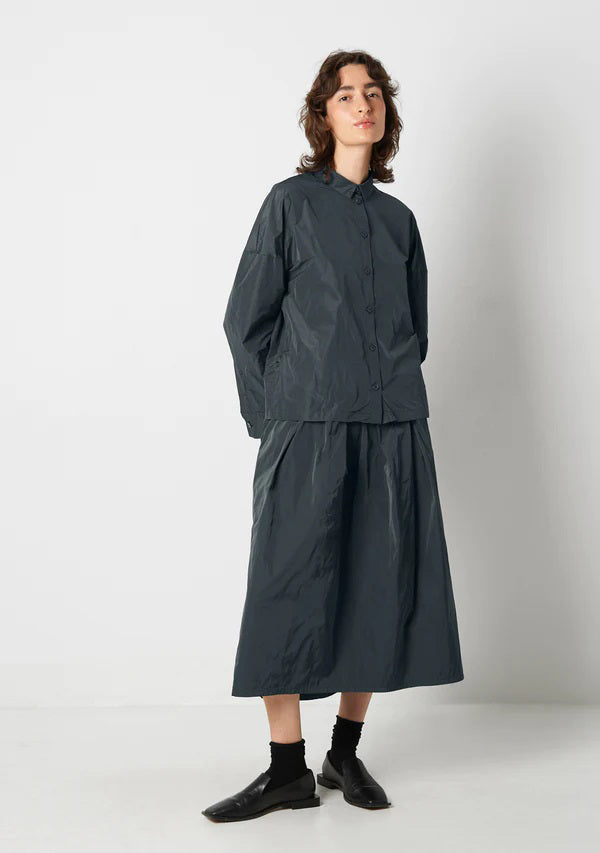 A person with shoulder-length wavy hair is wearing the KATHARINA HOVMAN Kimono Blouse from the brand KATHARINA HOVMAN. The blouse is long-sleeved, button-up, and dark gray. They are standing against a plain light gray background, gazing slightly upward with a calm expression while lightly clasping their hands in front.