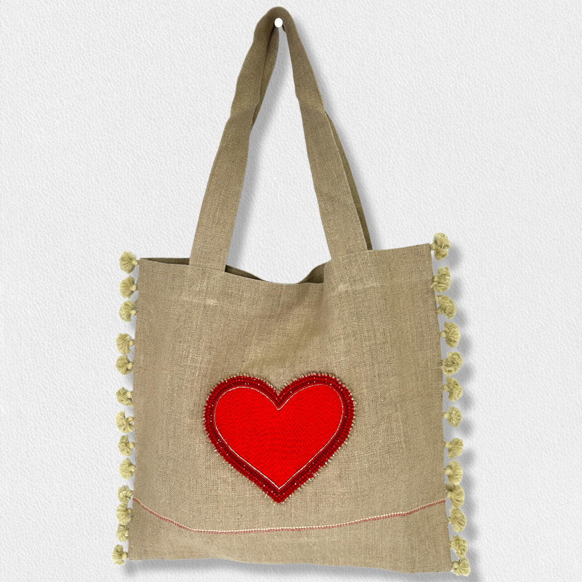 The LINEN MARKET TOTE from CITRUS FASHIONS is a charming brown linen tote with thin shoulder straps, featuring a large red heart design in the center and small yellow pompoms adorning both side seams. Hand-stitched with care, this delightful market bag hangs from a white textured wall by one of its straps.