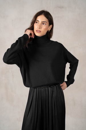 A woman with long brown hair smiles against a white background, wearing a ROSE & CROWN - CASHMERE TURTLENECK and wide-legged green pants, accessorized with necklaces, bracelets, and a large black handbag.