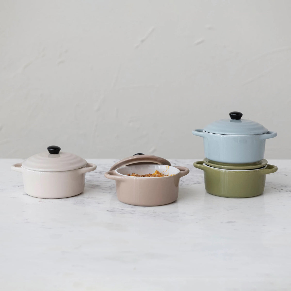 Four &quot;1 CUP MINI BAKER WITH LID&quot; casserole dishes by CREATIVE COOP are arranged in a 2x2 grid. The top left is light blue, the top right is olive green, the bottom left is light brown, and the bottom right is beige. Each piece of this versatile bakeware features a black knob on its lid.