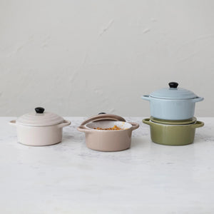 Four "1 CUP MINI BAKER WITH LID" casserole dishes by CREATIVE COOP are arranged in a 2x2 grid. The top left is light blue, the top right is olive green, the bottom left is light brown, and the bottom right is beige. Each piece of this versatile bakeware features a black knob on its lid.