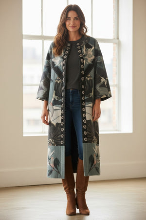 A woman stands indoors in front of a large window, smiling as she showcases the elegance of her FRANCO FERRARI - REVERSIBLE WOOL COAT. The long coat features a beautiful botanical floral print and is layered over a dark top, blue jeans, and brown knee-high boots.