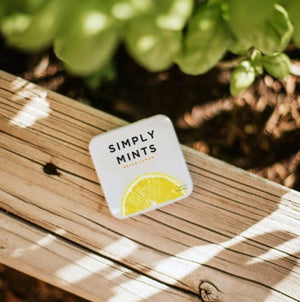A square tin container of Meyer Lemon Mints from Simply Gum is shown. The lid features the product name "Simply Gum" in large black letters with "Meyer Lemon Mints" in smaller yellow letters beneath it. A vibrant yellow lemon slice is depicted at the bottom, and there are no artificial sweeteners.