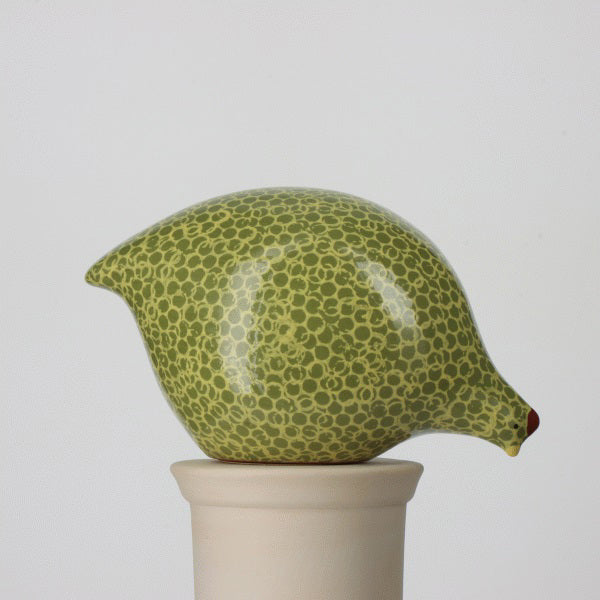 The LES CERAMIQUES DE LUSSAN - LARGE PECKING CERAMIC FRENCH GUINEA IN GREEN SPOTTED YELLOW sculpture, handmade in France by LES CERAMIQUES DE LUSSAN, resembles a plump bird akin to French guinea hens. It features a speckled green surface and is perched atop a cylindrical pedestal, with its glossy texture contrasted against a plain white background.