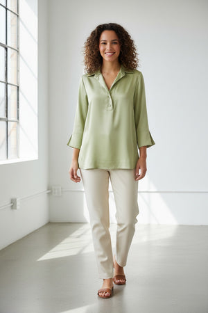 A woman with long brown hair is wearing the HINSON WU - AILEEN 3/4 Sleeve Liquid Satin Shirt in Pistachio, a light green collared blouse with three-quarter sleeves, paired with blue jeans as she stands against a plain, light-colored background. The shirt by HINSON WU features an elegant drape and luminous sheen, adding a touch of sophistication to her relaxed look.
