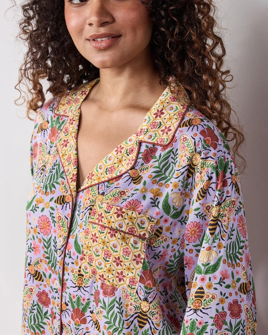 A person wearing the PRINTFRESH - BEE TAPESTRY ECO SATIN SOHO SET IN LAVENDER, featuring vintage-inspired bee and floral vine prints, holding a flower behind their back in a garden.