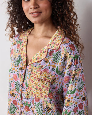 A person wearing the PRINTFRESH - BEE TAPESTRY ECO SATIN SOHO SET IN LAVENDER, featuring vintage-inspired bee and floral vine prints, holding a flower behind their back in a garden.