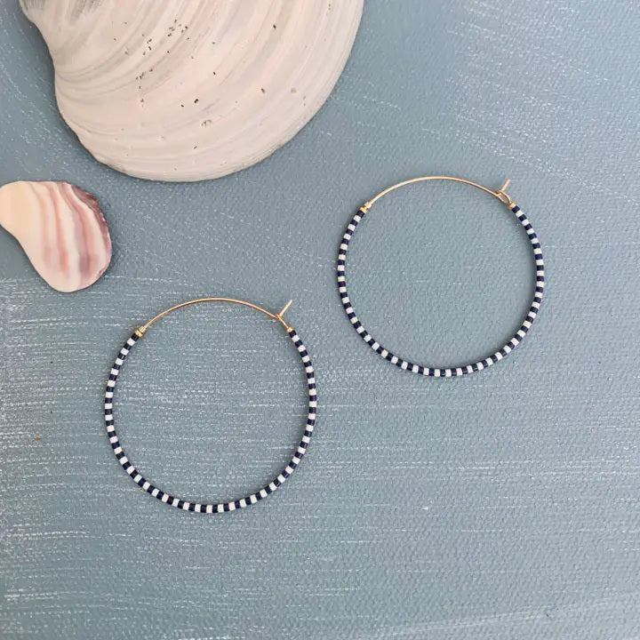 The ALICE RISE - BIG SINGLE NOTE BEADED HOOP EARRINGS feature gold-filled hoops with alternating black and white Japanese glass beads, displayed on a textured blue surface beside two seashells.